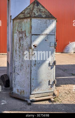 A weathered, hexagonal metal electrical cabinet or utility box with a pyramidal roof stands outdoors on a concrete surface. The structure is painted Stock Photo
