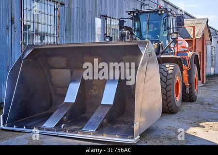 A large orange wheel loader with a heavy-duty front bucket attachment positioned on a concrete surface next to an industrial corrugated metal Stock Photo