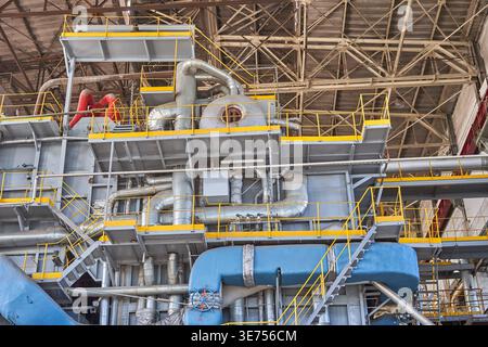 Interior view of a large industrial facility featuring complex heavy machinery and extensive piping systems. Multiple metal platforms with bright Stock Photo