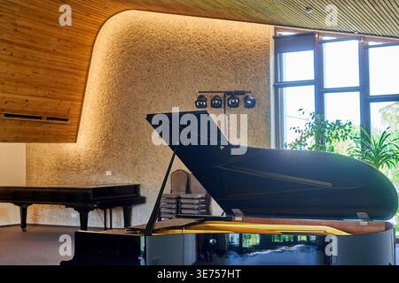 A glossy black grand piano with its lid open sits prominently in the foreground of a modern concert hall or performance space. The interior features Stock Photo