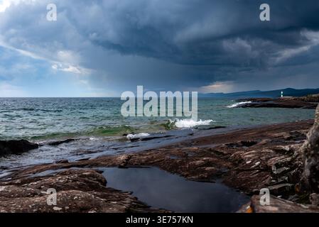 Dramatic Storm Over Rocky Coastline With Lighthouse and Rolling Waves Under Dark Clouds Stock Photo