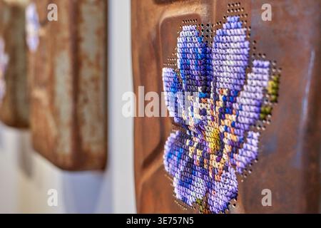 Close-up photograph of contemporary textile artwork featuring cross-stitch embroidery of a purple flower stitched onto a rusted metal surface. The Stock Photo