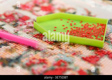 A green tray holds small red beads used for crafting. A pink stylus rests beside it on a colorful patterned fabric. Scene showcases a creative hobby Stock Photo