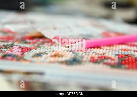 Close-up of a colorful diamond painting in progress. A pink applicator tool is used to place sparkling gems on a floral design Stock Photo