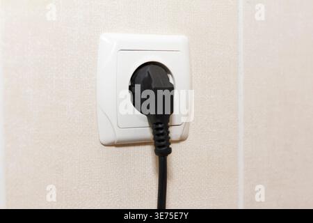 A close-up of a black electrical plug inserted into a white wall socket. Background is a light-colored wall, emphasizing plug and socket Stock Photo