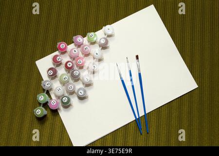 Art supplies arranged on a textured green background. Includes paint pots in various colors, three paintbrushes, and a blank canvas Stock Photo