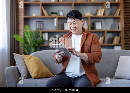 Asian man sitting comfortably on a gray sofa at home, smiling while engaging with a tablet computer, representing connection, leisure, and modern digital lifestyle Stock Photo