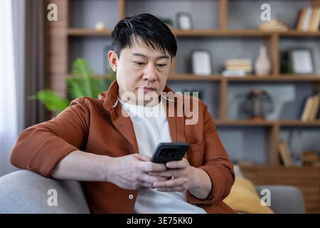 Asian man sitting comfortably on a sofa, looking intently at his smartphone, connecting, communicating, and interacting with technology in his modern home environment Stock Photo
