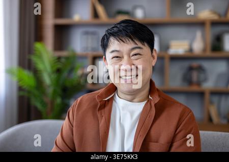 Smiling mature asian man wearing casual clothes, feeling happy and confident while relaxing in his modern home living room with bookshelves behind him Stock Photo