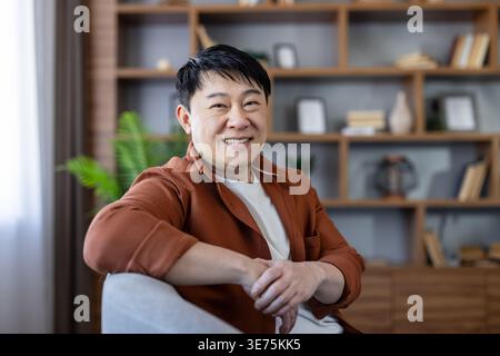 Smiling asian man looking at camera sitting in a comfortable armchair, feeling happy and relaxed in his modern living room with a bookshelf in the background Stock Photo
