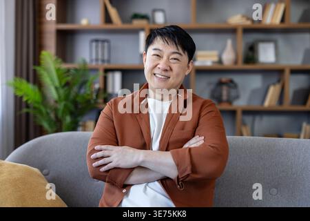 Mature asian man smiling confidently with arms crossed while seated on a modern living room sofa, relaxed and self-assured, conveying domestic comfort, success and wellbeing Stock Photo