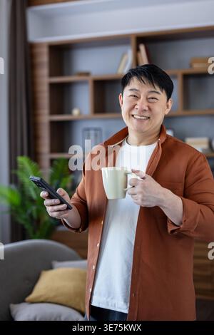 Mature asian man smiling at camera while holding smartphone and coffee cup in a cozy living room, enjoying a relaxed morning of browsing, messaging and everyday contentment Stock Photo
