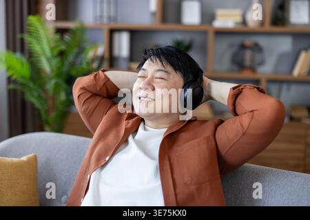 Asian man enjoying favorite audio, closing eyes with hands behind head, relaxing on a sofa at home, appreciating leisure time and finding calm in his comfortable living room Stock Photo