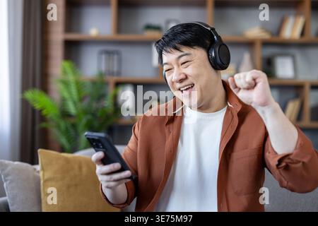 Young asian man wearing headphones and holding a smartphone, celebrating a moment of victory or success at home, expressing happiness and joy while relaxing on a couch Stock Photo