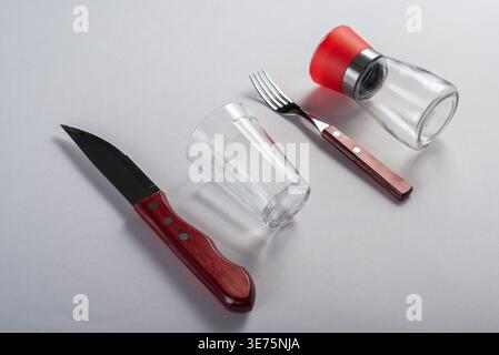 Isolated on white. Minimalist setup featuring knife, fork, glass and pepper mill. Soft diffused lighting emphasizes wood textures and clear glass. Com Stock Photo