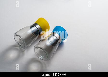Two salt and pepper grinders in minimalist setup, isolated on white. Reflective glass, stainless steel, yellow and blue plastic tops. Food and lifesty Stock Photo