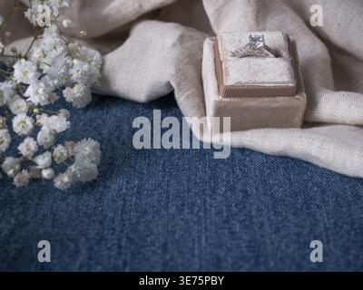 Engagement ring displayed in velvet ring box on rustic linen and denim with delicate white flowers creating romantic western wedding proposal Stock Photo
