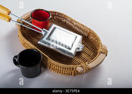 Studio shot features waffle iron, red and black mugs in woven basket, against neutral backdrop. Soft lighting accentuates metallic and textile texture Stock Photo