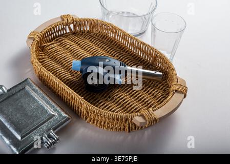 Studio shot featuring culinary torch nestled inside wicker basket. Clear glasses add simplicity. High key lighting reveals textures of metal, glass an Stock Photo