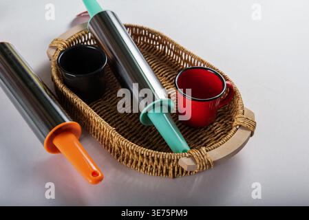 Two rolling pins, black and red cups arranged inside wicker basket, neutral background. Shiny metal, vibrant color handles. Kitchenware concept for ba Stock Photo