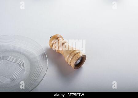Minimalist setup featuring wooden pepper mill adjacent clear glass plate on clean white surface. Gentle lighting highlights smooth wood grain, subtle Stock Photo