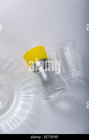 Overhead shot features glass plate, glass cup, and transparent glass container with yellow plastic top on white. Soft studio lighting, minimal shadows Stock Photo
