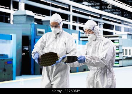 Engineering team processing silicon wafers in automated manufacturing plant. Teamworking colleagues inspecting delicate semiconductor substrates hardware during quality control process Stock Photo