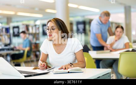Positive Latin young lady in casual clothes and glasses using a computer in a quiet library Stock Photo