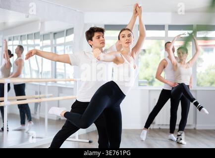 Pretty female ballerina and male partner practicing ballet arabesque pose during training with group active people in dancing hall Stock Photo