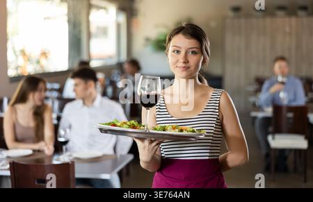 Waitress standing with salad and glass of wine on serving tray in restaurant Stock Photo
