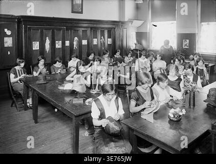 Historical image of a girls’ millinery design class at the East Side Continuation School in New York City, with students sewing and shaping hats at long worktables under classroom instruction. Photograph by Bain News Service. Stock Photo