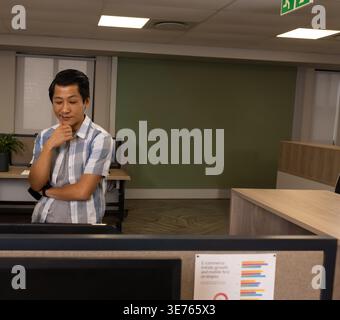 Mid adult Asian man leaning behind divider at office viewing monitor, pinned chart, copy space. Workspace, contemplation, professional, modern, lighti Stock Photo