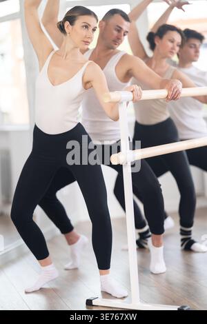 Group of sports people standing in row together while doing ballet dance moves demi plie while holding on ballet barre in ballet studio Stock Photo