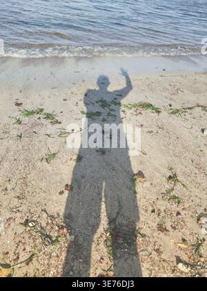 Human shadow casting on the sand and water edge, reflecting a person waving Stock Photo