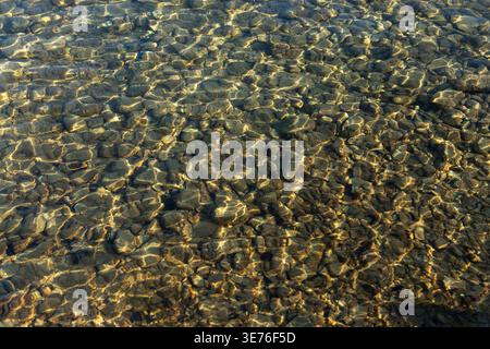 Sunlit clear water ripples over smooth pebbles, creating a natural abstract texture background with blue and golden tones. Stock Photo