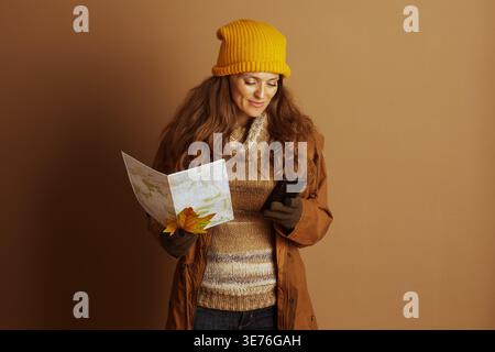 A woman in autumn attire, wearing a yellow knit hat and brown coat, looks down, navigating with a smartphone and open map, embodying modern outdoor ex Stock Photo