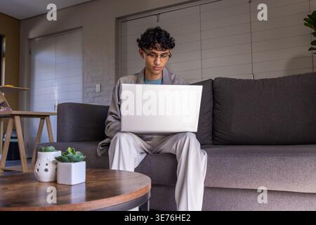 Indian man sitting on gray sofa at home, wearing blazer and clear glasses using silver laptop Stock Photo
