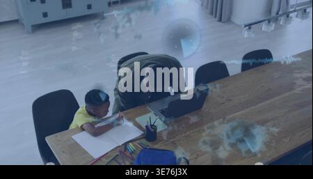 Leaning olive-jacketed man helping yellow-shirted boy at dining table, using laptop and pencils Stock Photo