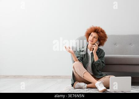 Redhead female freelancer with laptop talking by mobile phone near light wall Stock Photo