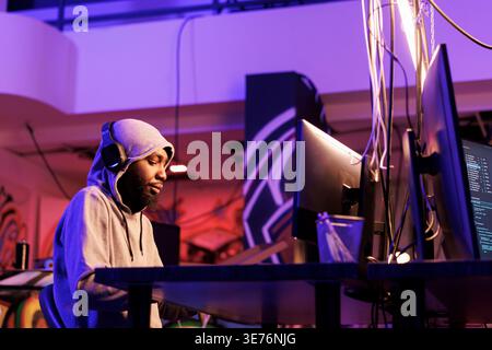 African american hacker wearing headphones and coding malicious software for cyberattack. Man in hood listening to music in earphones while breaching online server at night Stock Photo