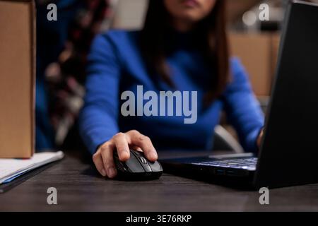 Woman managing parcel delivery on laptop in retail ecommerce warehouse. Logistics manager doing inventory check on laptop while using computer mouse with close up on hand Stock Photo