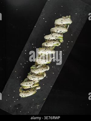 Spiral sushi rolls on dark slate plate. White sauce drizzled over green avocado. Black sesame seeds scattered for texture. Minimalist black background Stock Photo
