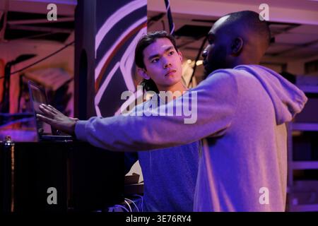 Hackers discussing malware code on laptop while planning illegal activities. African american and asian criminals talking about online server hacking technique in abandoned warehouse Stock Photo