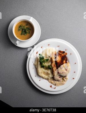 Cozy meal served on minimalist gray table. Golden soup garnished with fresh herbs beside plate. Mashed potatoes and grilled steak topped with creamy s Stock Photo