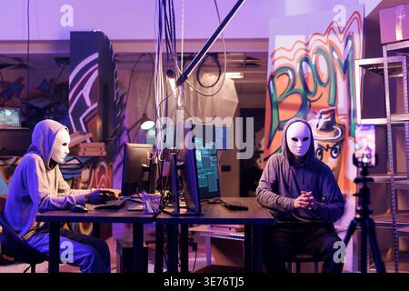 Internet scammers in anonymous masks threatening victim online on smartphone. Hackers recording ransom message video on mobile phone while working together in abandoned warehouse Stock Photo