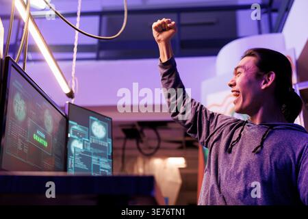 Happy hacker making victory gesture after getting server access granted. Cheerful excited asian man celebrating hacking computer network system successfully and raising fist Stock Photo