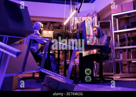 Two hackers coding computer worm on multiple computer screens in dark abandoned warehouse. Young men in hoods hacking online server and stealing privacy information from database Stock Photo