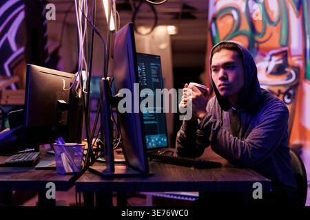 Hacker drinking coffee to go while doing illegal criminal activity on computer. Young man in hood holding beverage mug and doing ddos attack on website in abandoned warehouse Stock Photo