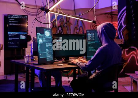 Hacker in hood using spyware to steal information from server while coding on computer with multiple screens. Man programming illegal malicious software in hideout place Stock Photo