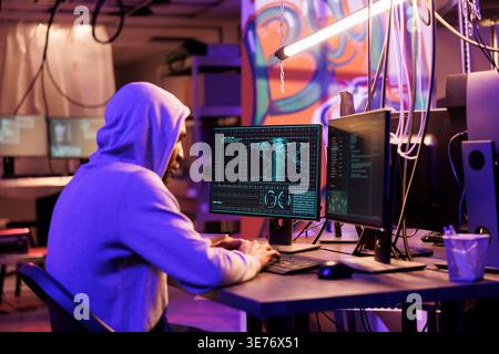 African american hacker in hood breaking into server and stealing sensitive information. Internet criminal coding malicious software for data breach on computer in dark abandoned warehouse Stock Photo